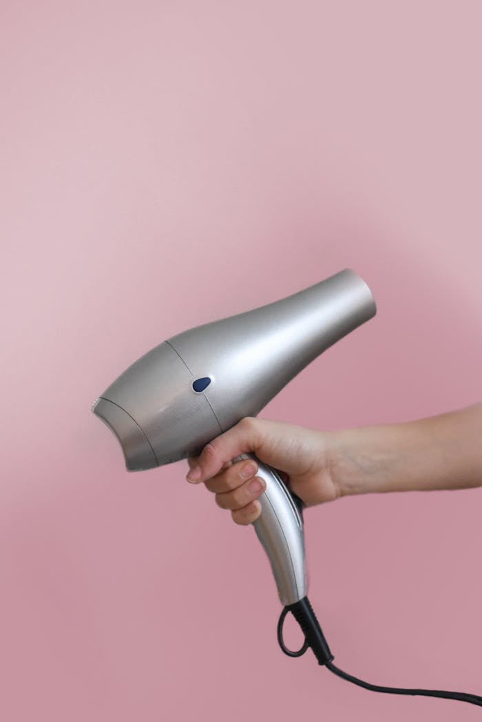Close-up of a hand holding a silver hair dryer on a pink background.